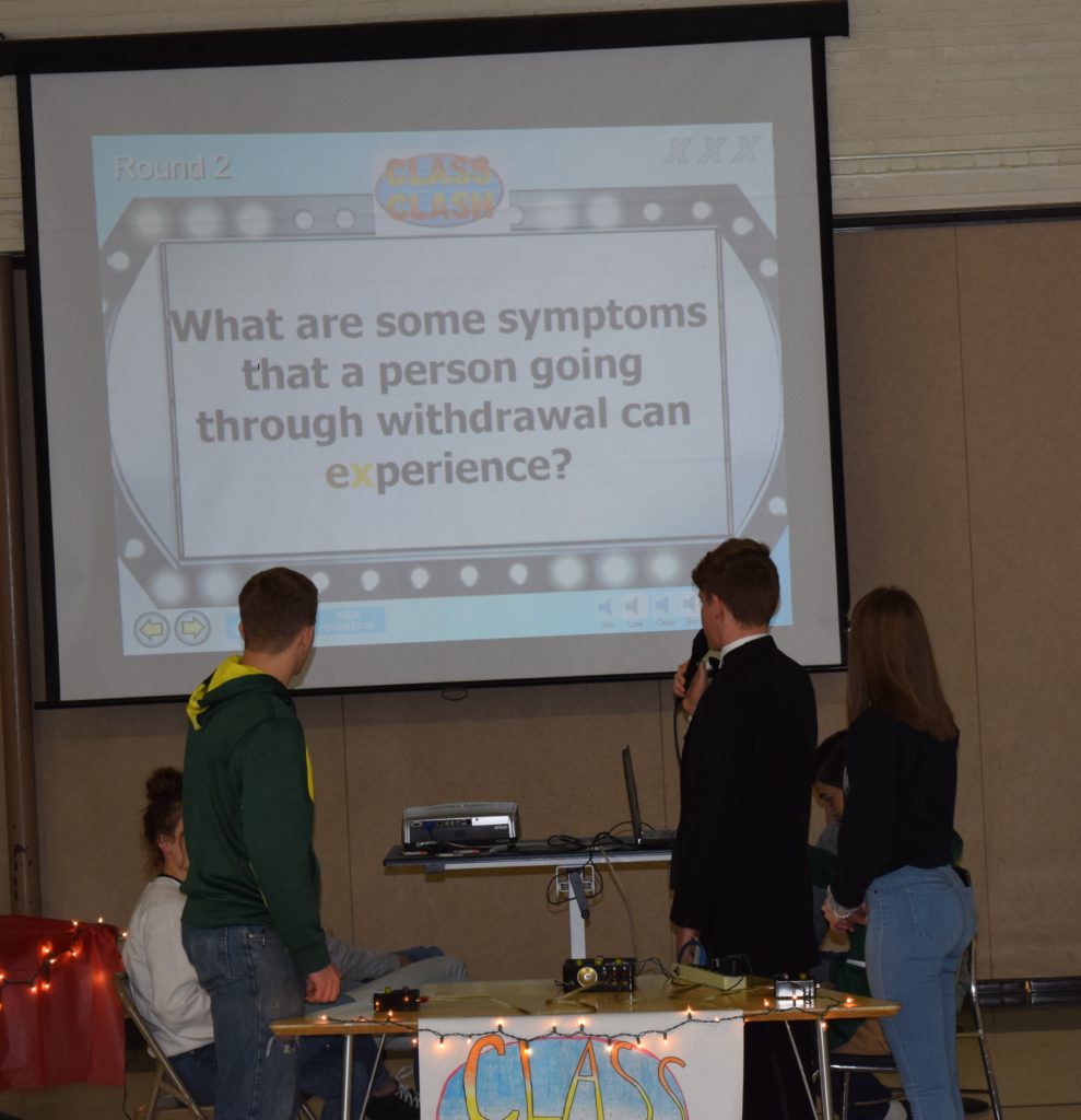 Two teams of students stand before a projection screen. The screen shows the question "What are some symptoms that a person going through withdrawal can experience?" A poster proclaims "Class Clash."